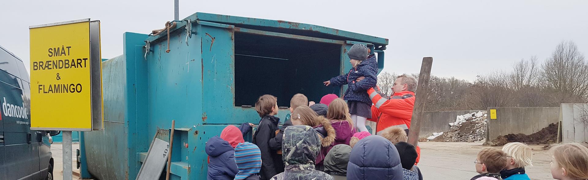 Genbrugsvejleder løfter barn op til container mens andre børn kigger på