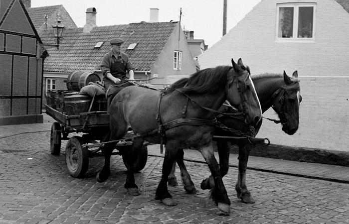 Gammelt sort/hvid foto af mand der bliver trukket i hestevogn af to heste gennem gade med rendesten