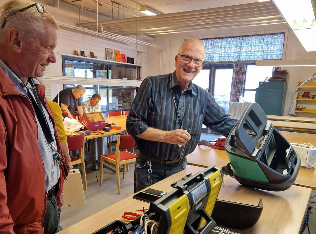 To mænd smiler og står ved bord, i færd med at reparere en støvsuger