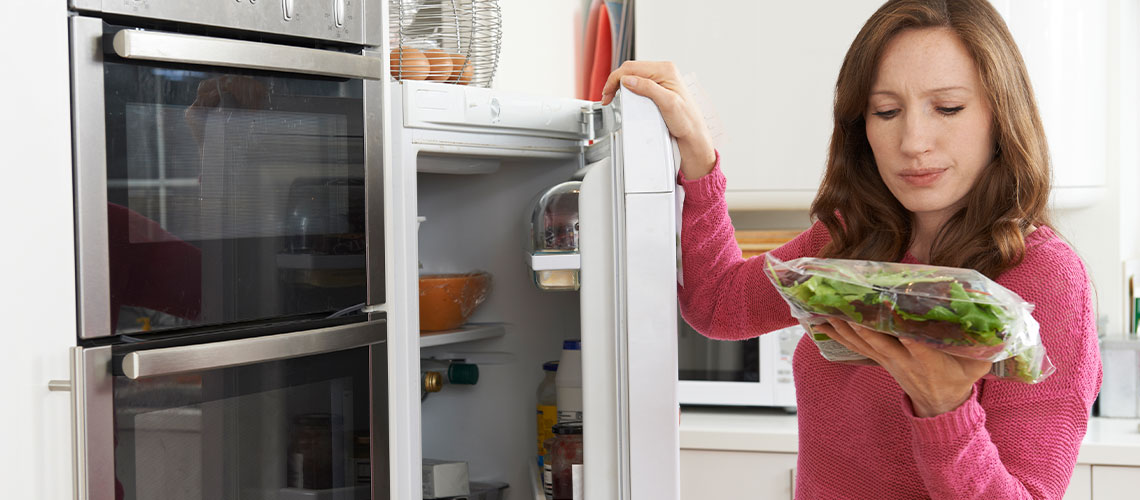 Kvinde står ved køleskab med en pose salat i hånden og kigger lidt skeptisk på posen
