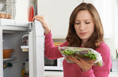 Kvinde står ved et åbent køleskab med en pose salat. Hun kigger skeptisk på posen.