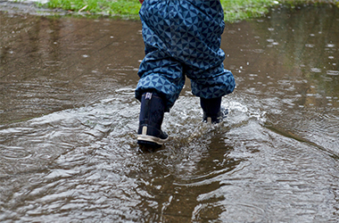 Barn i blåt regntøj og gummistøvler går i stor vandpytte